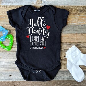 May include: Black baby bodysuit with white script text reading "Hello Daddy I CAN'T WAIT TO MEET YOU! January 2025", with red heart details. A blue and green teether and white baby socks are also in the image.