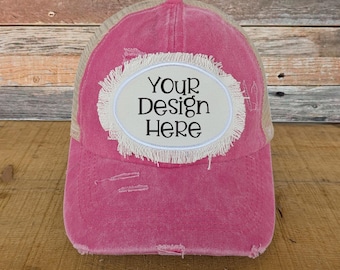 May include: Pink distressed trucker hat with a beige mesh back. The front features an oval patch with the text "Your Design Here" in black. The hat has a pre-curved bill and a distressed look.