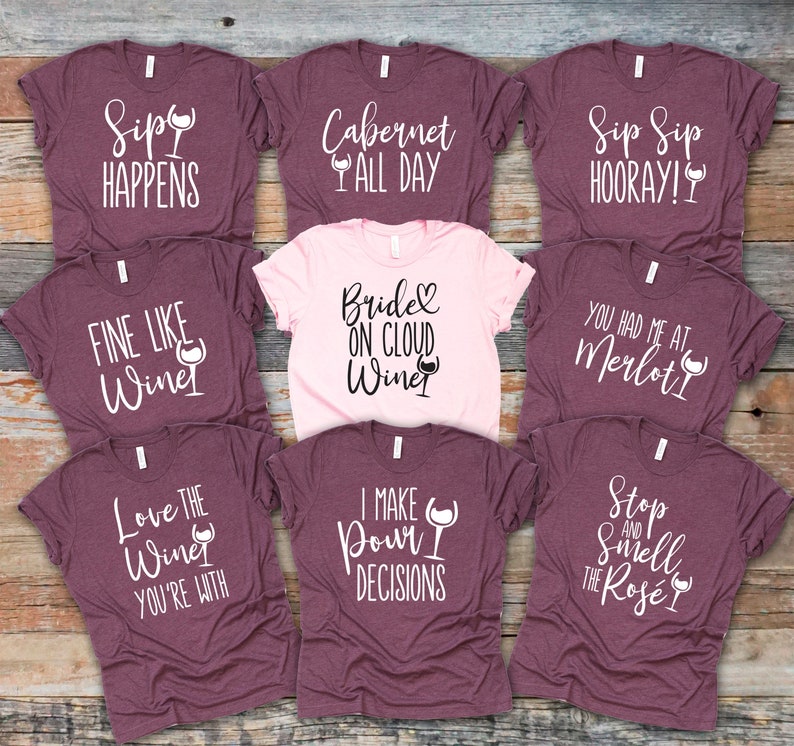 May include: Nine different wine-themed t-shirts in various shades of purple and pink. The shirts feature a variety of wine-related puns and sayings, such as "Sip Sip Hooray!" and "Fine Like Wine".
