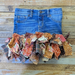 Puede incluir: Unos pantalones cortos de mezclilla azul con cintura con trabillas y cierre de botón metálico. El dobladillo está adornado con un fleco denso y superpuesto de retazos de tela variados en tonos marrón, naranja, crema y estampados florales.
