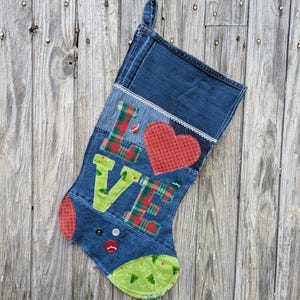 Op de afbeelding: Een kerstkous van spijkerstof met het woord LOVE in groen en rood ruitjespatroon, waarbij een rood hart de letter O vervangt. De kous is voorzien van witte kanten randen, een groen patroon bij de teen en drie knopen bij de hiel.