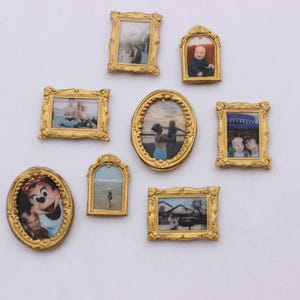 Puede incluir: Una colección de ocho marcos de fotos dorados y ornamentados de varias formas y tamaños, cada uno con una fotografía. Los marcos están dispuestos en una pared blanca, mostrando fotos familiares y retratos. Los marcos varían en tamaño y forma.