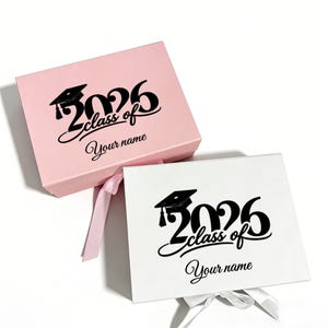 Puede incluir: Dos cajas de regalo de graduación, una rosa y otra blanca, con texto negro. El texto de cada caja dice "2026 class of" y "Your name" con un gráfico de birrete. Cada caja tiene un cierre de cinta.
