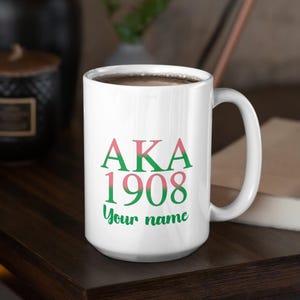 Puede incluir: Taza de cerámica blanca con el texto "AKA 1908" en verde y rosa. Las palabras "Your name" están debajo de los números. La taza tiene un asa curva y está sobre una superficie de madera oscura.