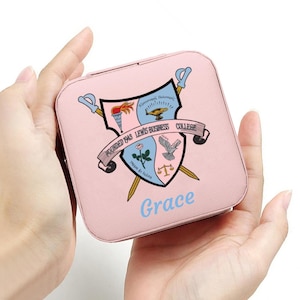 May include: A pink square jewellery box featuring a shield design and the words "Founded 1943 Lewis Business College" on a banner. The name "Grace" is written in light blue. Two swords cross behind the shield.