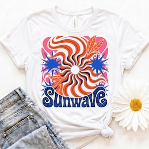 May include: White t-shirt with a vibrant graphic. The design includes an orange and white striped sunburst, blue star shapes, and the word "SUNWAVE" in blue. The shirt is shown with blue denim jeans.