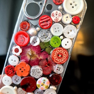 May include: A clear phone case decorated with a dense arrangement of assorted buttons in various colours, sizes, and shapes, including red, white, orange, and green. Pressed dried flowers in shades of pink and red are embedded among the buttons.