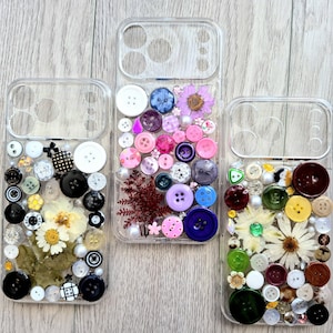 May include: Three clear plastic phone cases decorated with various colorful buttons, small white pearls, and pressed dried flowers. The buttons vary in size and color, including black, white, purple, pink, green, and brown, arranged in a dense collage.