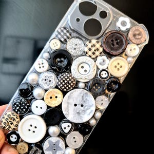 May include: A clear smartphone case decorated with a dense collage of various buttons in black, white, grey, and beige. The buttons feature diverse textures, patterns, and shapes, including checkered, marbled, star-shaped, and floral designs.