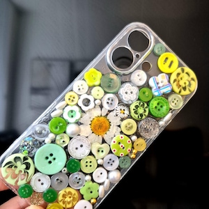May include: A clear phone case decorated with a collage of various buttons in shades of green, yellow, and white, featuring a central pressed daisy flower, small metallic beads, and circular cutouts for a camera lens and flash on the upper right.