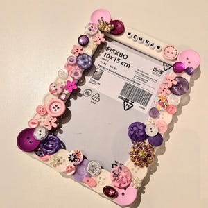 May include: A decorative photo frame, embellished with an assortment of pink, purple, and white buttons, beads, and floral accents. The frame is labeled "FISKBO 10x15 cm (4x6")" and the name "EMMA" is spelled out on the top.