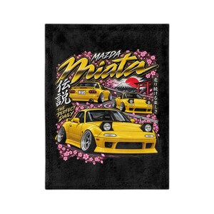 May include: A black blanket featuring a graphic illustration of three yellow Mazda Miata sports cars on a winding road. The design includes pink cherry blossoms, a red sun, a mountain silhouette, and text reading Mazda Miata, The Perfect Daily.