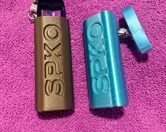 Op de afbeelding: Twee beschermhoezen voor een vape-apparaat, één goudkleurig en één blauw, beide met de tekst "SPKO" in reliëf. De gouden hoes bevat een zwart vape-apparaat. Een lichtblauwe lanyard is bevestigd aan de blauwe hoes. De hoezen liggen op een paars oppervlak.