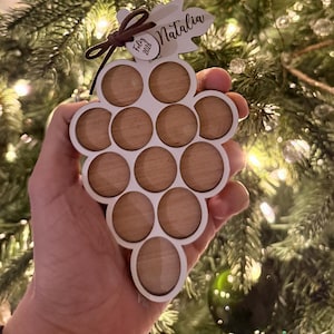 Puede incluir: Adorno navideño de madera con forma de uva, con borde blanco y círculos marrones. El adorno tiene una etiqueta que dice "Natalia" y "Feliz 2023". Se sostiene frente a un árbol de Navidad con luces y otros adornos.
