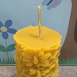May include: A yellow beeswax candle with a wick, cylindrical in shape. The candle is decorated with a honeycomb pattern and floral and bee designs. The background features a light blue wall with a butterfly and owl illustration.