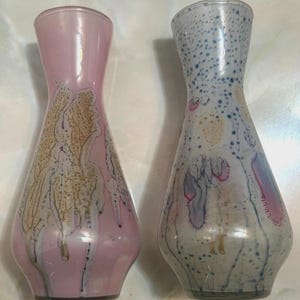 Puede incluir: Dos jarrones decorativos de cristal. Uno es rosa con un patrón blanco y dorado, el otro es blanco con un patrón azul y morado. Ambos tienen una parte superior acampanada y una base bulbosa.