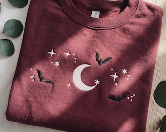 May include: A maroon crewneck sweatshirt featuring white embroidered celestial motifs including a crescent moon, various stars, and three black bats. The embroidery is centered on the chest of the garment, which has a ribbed collar and long sleeves.