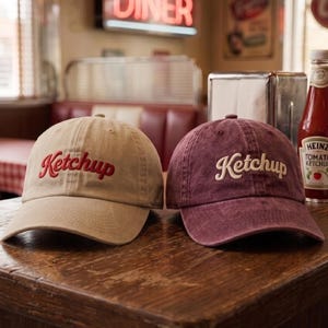 May include: Two baseball caps sit side by side. The left cap is tan with red embroidered text that reads Ketchup. The right cap is a faded maroon color with white embroidered text that reads Ketchup. Both caps feature a curved brim and a structured crown.