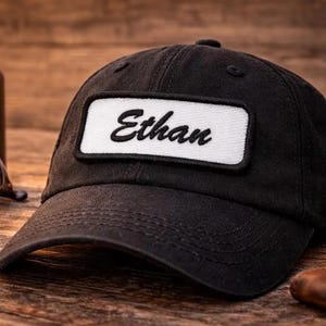 May include: Black baseball cap with a curved brim. The cap features a white rectangular patch with the name "Ethan" embroidered in black script. The cap is sitting on a wooden surface.