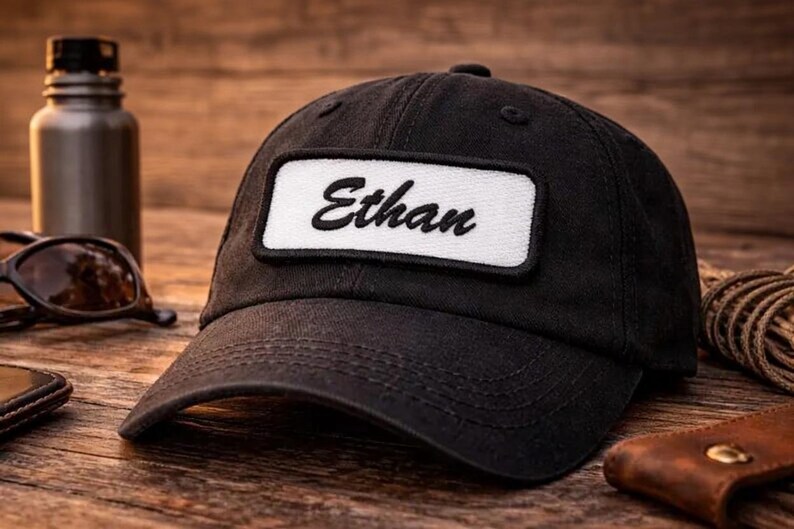 May include: Black baseball cap with a curved brim. The cap features a white rectangular patch with the name "Ethan" embroidered in black script. The cap is sitting on a wooden surface.