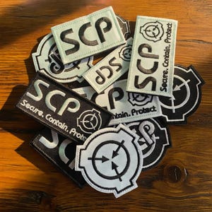 May include: A collection of embroidered patches in various shapes and sizes. The patches feature the letters "SCP" in white or black, along with the words "Secure, Contain, Protect" and a circular logo. The patches are in shades of black, white, and light green.