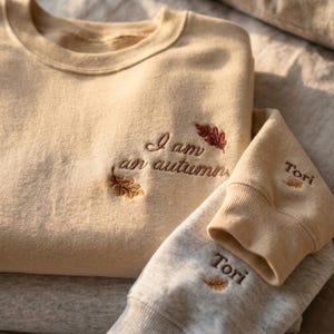 May include: A beige crewneck sweatshirt featuring brown embroidered text that reads I am an autumn with two small embroidered leaves. The sleeve cuff displays the name Tori in brown embroidery accompanied by a tiny leaf icon.