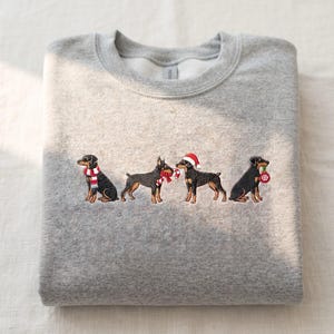 May include: A heather gray crewneck sweatshirt featuring an embroidered design of four black and tan dogs. The dogs are decorated with festive items including a red and white striped scarf, a candy cane, a Santa hat, and a red ornament.