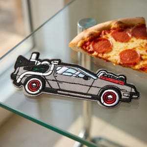 May include: Embroidered patch featuring a gray DeLorean car with red and white accents. The patch is on a glass surface next to a slice of pizza with pepperoni. The car patch is a fun accessory.