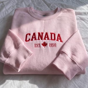 May include: A light pink crewneck sweatshirt featuring red embroidered text that reads CANADA, with the text EST 1950 flanking a small red maple leaf icon centered below the main word. The garment has long sleeves with ribbed cuffs.