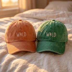 May include: Two baseball caps, one in a faded orange and one in a faded green, each featuring the word WILD embroidered in white thread on the front panel, with a small pine tree icon replacing the letter I in the word.