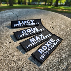 May include: Four black embroidered patches with white text. The patches display the names "LUCY", "ODIN", "MAX", and "ROXIE", each followed by the phone number (973)-123-9872. The patches are rectangular and have a stitched border.
