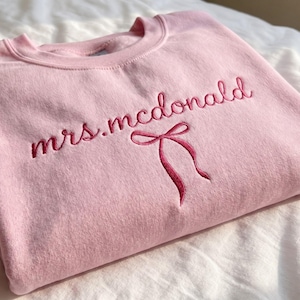 May include: A light pink crewneck sweatshirt featuring the embroidered text "mrs.mcdonald" in a dark pink cursive font, positioned above a small, matching dark pink embroidered bow graphic centered on the chest of the garment.