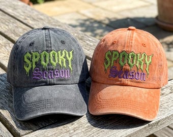 May include: Two baseball caps, one in charcoal gray and one in orange, both featuring a distressed fabric finish. Each cap has embroidered text that reads "SPOOKY" in a dripping lime green font above the word "Season" in a purple script font.