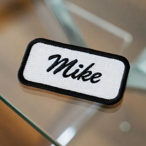 May include: A rectangular white patch with the name "Mike" embroidered in black script. The patch has a black border and is likely designed to be sewn onto clothing or accessories. The patch is on a glass surface.
