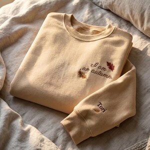 May include: A beige crewneck sweatshirt featuring embroidered text that reads I am an autumn. The design includes two small embroidered leaves near the text. The sleeve features the embroidered name Tori with a small leaf icon below it.