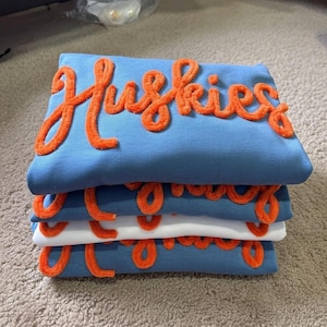 May include: A stack of four folded sweatshirts, three in light blue and one in white, each featuring the word Huskies in large, raised, orange chenille-style script lettering across the front, creating a textured and layered appearance.