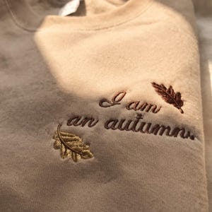 May include: A beige crewneck sweatshirt featuring dark brown embroidered text that reads I am an autumn. The embroidery is accented by two small leaf designs, one in a golden yellow tone and the other in a deep brown shade.