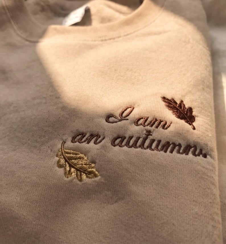 May include: A beige crewneck sweatshirt featuring dark brown embroidered text that reads I am an autumn. The embroidery is accented by two small leaf designs, one in a golden yellow tone and the other in a deep brown shade.