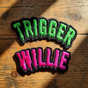 May include: Two embroidered patches with black borders. The top patch reads "TRIGGER" in green with dripping accents. The bottom patch says "WILLIE" in pink with dripping accents. Both patches are on a wooden surface.