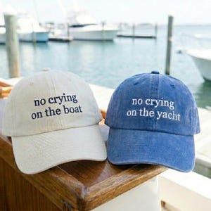 May include: Two baseball caps with embroidered text. The off-white cap features the black text 'no crying on the boat' and the blue cap features the white text 'no crying on the yacht'. Both caps have a curved brim and a structured crown design.