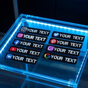 May include: Black rectangular stickers with social media logos and the text "YOUR TEXT" in white. Logos include Instagram, Facebook, YouTube, VK, Twitter, Pinterest, Twitch, and Google. The stickers are on a clear, illuminated surface.