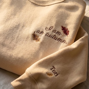 May include: A beige crewneck sweatshirt featuring embroidered text that reads I am an autumn. The design includes small brown and red leaf motifs near the text. The sleeve cuff features the embroidered name Tori with a small leaf icon.