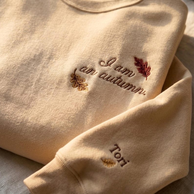 May include: A beige crewneck sweatshirt featuring embroidered text that reads I am an autumn. The design includes small brown and red leaf motifs near the text. The sleeve cuff features the embroidered name Tori with a small leaf icon.