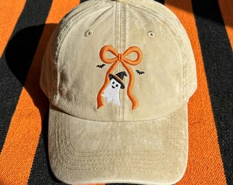 May include: A tan baseball cap featuring an embroidered design on the front. The embroidery depicts a white ghost wearing a black witch hat, positioned beneath a large orange ribbon bow, with two small black bats flanking the ghost.