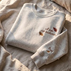 May include: A light heather gray crewneck sweatshirt featuring embroidered text on the chest that reads I am an autumn with two small oak leaves. The sleeve cuff includes the embroidered name Tori with a single small leaf icon.