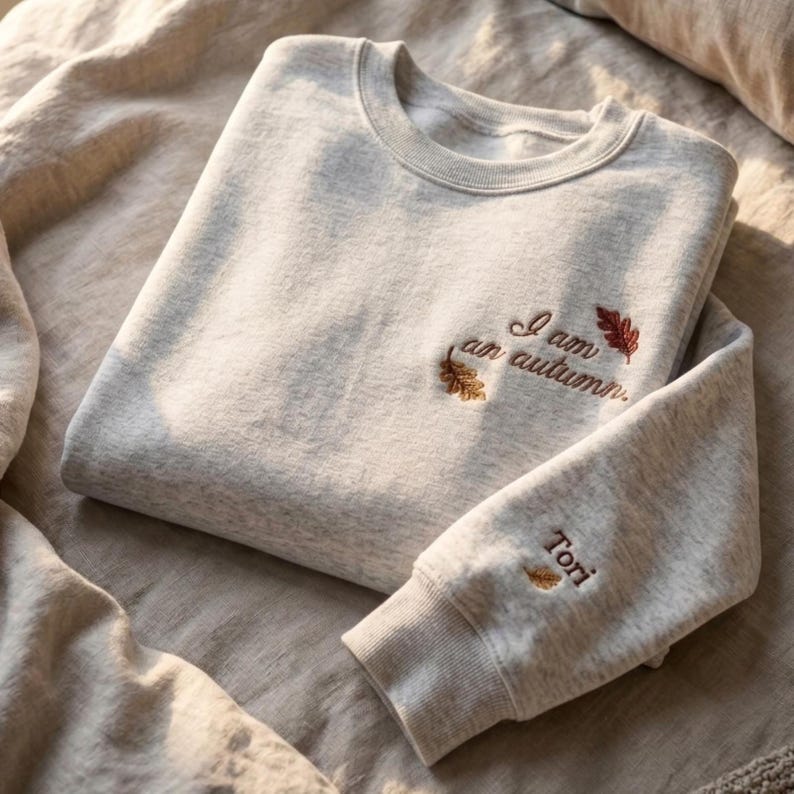 May include: A light heather gray crewneck sweatshirt featuring embroidered text on the chest that reads I am an autumn with two small oak leaves. The sleeve cuff includes the embroidered name Tori with a single small leaf icon.