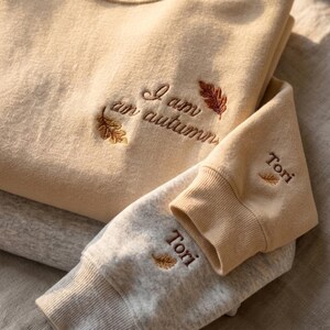 May include: A beige sweatshirt featuring the embroidered text I am autumn with two small leaf icons. The sleeve cuff is embroidered with the name Tori and a small leaf icon. A second gray sweatshirt is visible underneath with the name Tori.