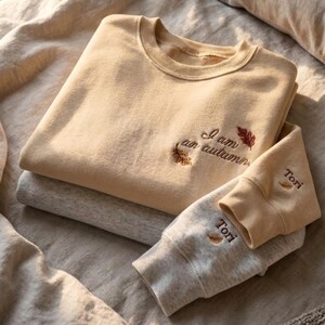 May include: A beige crewneck sweatshirt featuring the embroidered text I am an autumn with two small leaf icons. A second light gray sweatshirt is layered underneath, with the name Tori and a small leaf icon embroidered on the sleeve cuff.