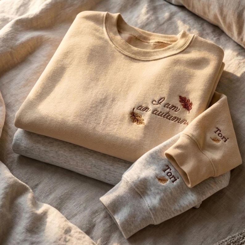 May include: A beige crewneck sweatshirt featuring the embroidered text I am an autumn with two small leaf icons. A second light gray sweatshirt is layered underneath, with the name Tori and a small leaf icon embroidered on the sleeve cuff.