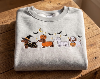 May include: A light gray crewneck sweatshirt featuring an embroidered design of four dachshunds in Halloween costumes. The dogs are dressed as a witch, a pumpkin, a mummy, and a ghost, surrounded by small embroidered bats, stars, and a crescent moon.